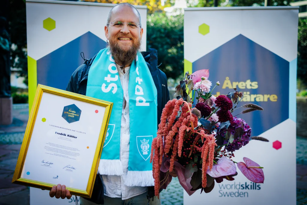 Man with beard smiling after winning regional VET teacher of the year. He has a sash and has flowers and a diploma in hand