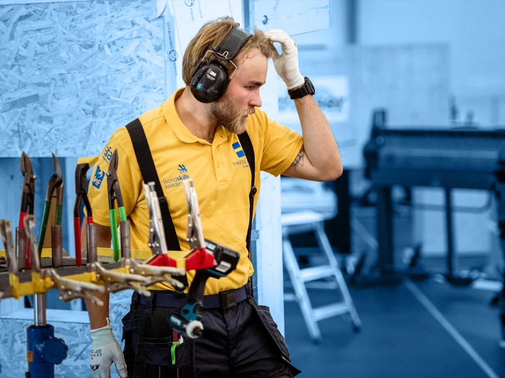 A young man who wear safety gear stands behind a tool rack looking to the right while brushing his hair aside with his hand