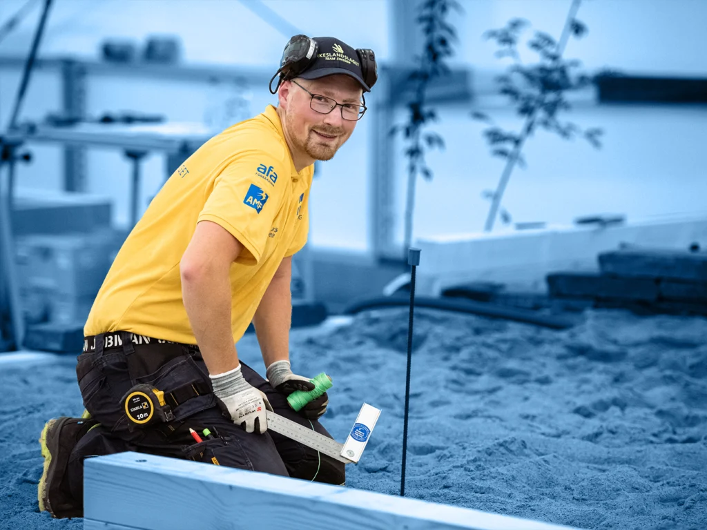 A young man sits on the ground wear work pants, ear protection and holding an angle hook